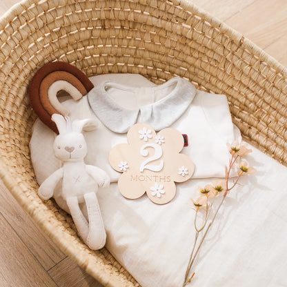 A picture of a crib with baby clothing, toys, flower, and a flower shaped wooden milestone card displaying 2 months