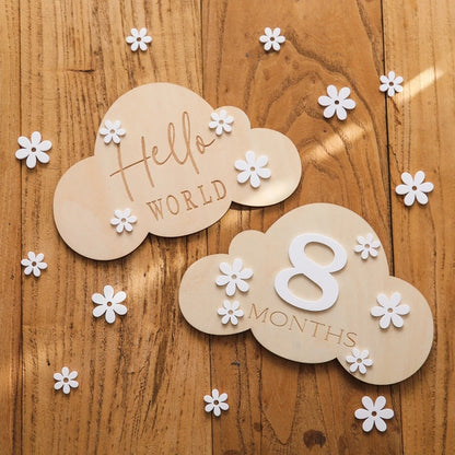 A cloud shaped wooden milestone card displaying Hello World alongside another card displaying 8 months, set against a wooden background with white flower cutouts
