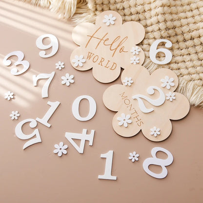 A flower shaped wooden milestone card displaying Hello World alongside another card displaying 2 months, in an aesthetic background with white flower and number cutouts