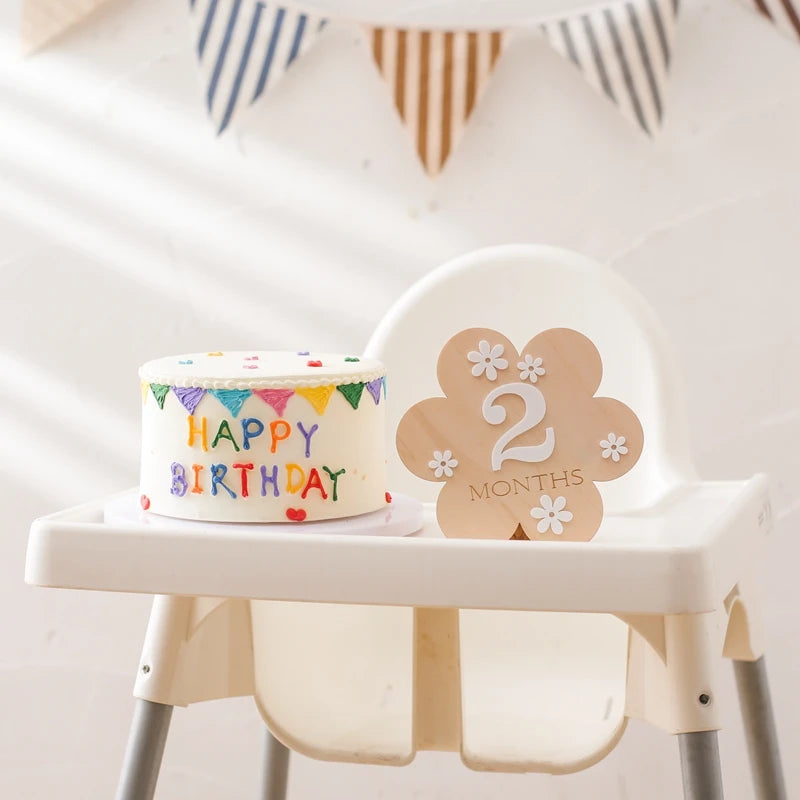 A flower shaped wooden milestone card displaying 2 months alongside a cake in a baby highchair and a party banner in the background