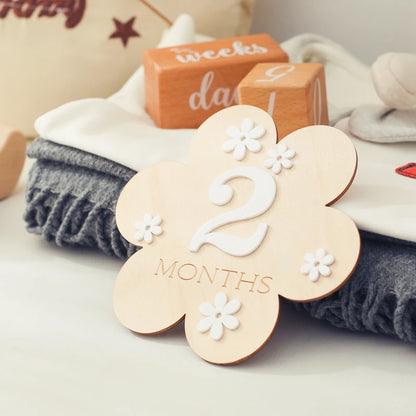 A flower shaped wooden milestone card displaying 2 months leaning beside folded clothes