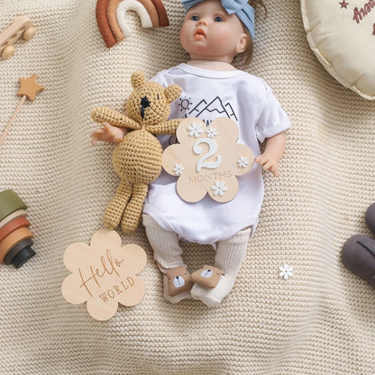 A flower shaped wooden milestone card displaying Hello World alongside a baby holding a card displaying 2 months and a teddy bear