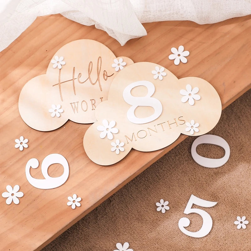 A cloud shaped wooden milestone card displaying Hello World alongside another card displaying 8 months, set against a wooden background with white flower and number cutouts
