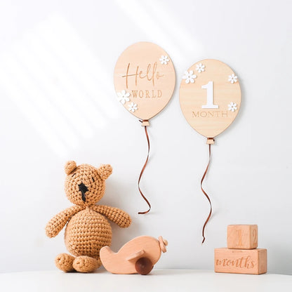 A balloon shaped wooden milestone card displaying Hello World alongside another card displaying 1 months, with bears, toys on a white wall background