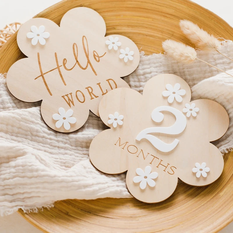 A flower shaped wooden milestone card displaying Hello World alongside another card displaying 2 months, in a wooden aesthetic backround